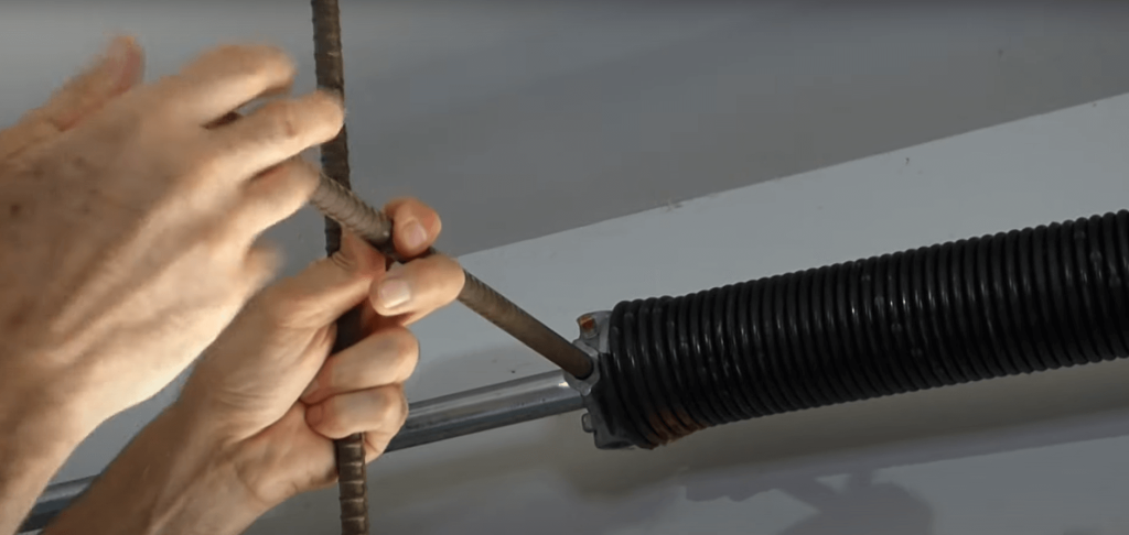 Close-up of a person adjusting the tension on a garage door torsion spring using winding bars. The black coil spring is mounted on a metal shaft, and the person’s hands are carefully inserting the bars into the winding cone.