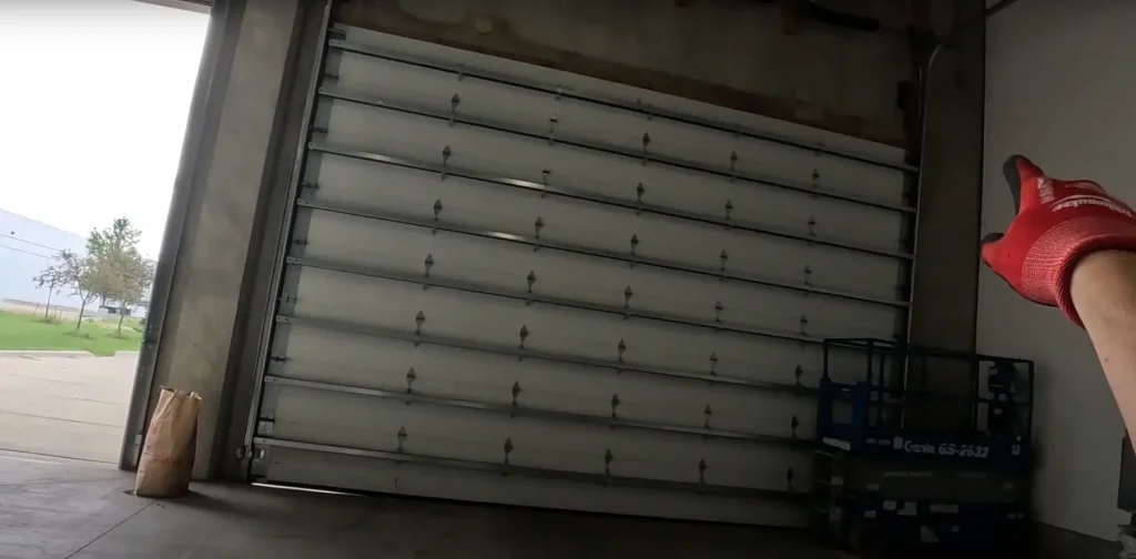 A worker wearing red gloves points at a large, misaligned commercial garage door inside a warehouse, indicating damage or an issue with its track or mechanism.