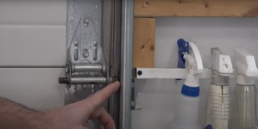 A person pointing at the hinge and roller mechanism of a garage door track, highlighting the importance of regular lubrication. Nearby, a row of spray bottles containing cleaning and maintenance solutions is mounted on the wall.