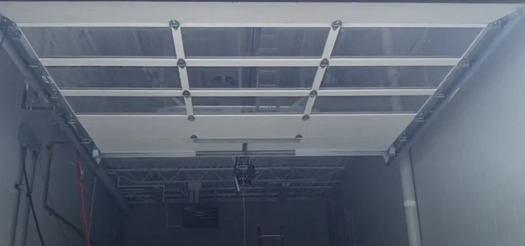 A commercial garage door with transparent panels partially open, revealing the interior ceiling structure and opener mechanism.