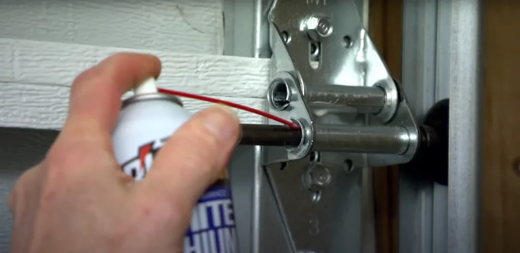 Hand applying lubricant to a commercial garage door hinge using a spray can with a red straw nozzle.