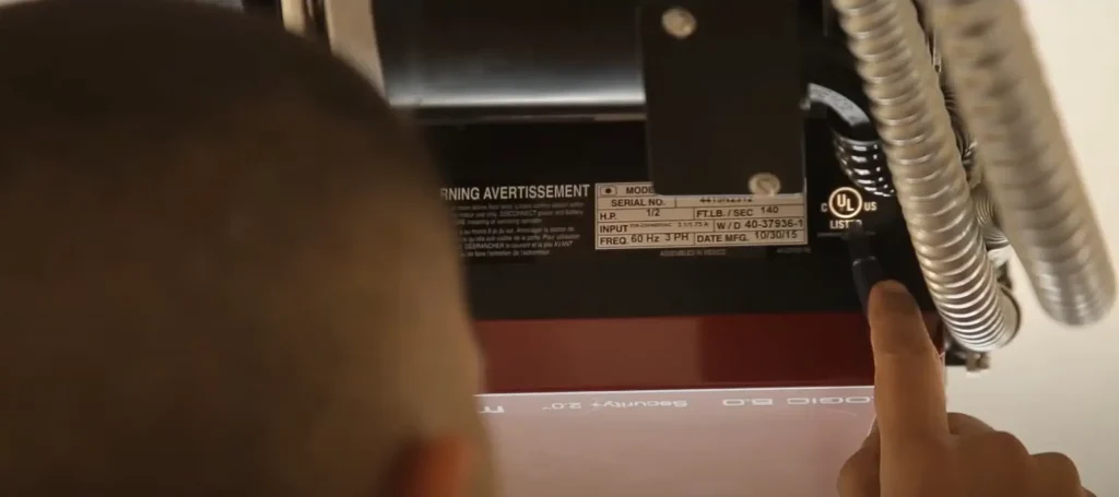 A person pointing to the UL certification label on a commercial garage door motor. The label includes specifications such as model number, serial number, and manufacturing date.