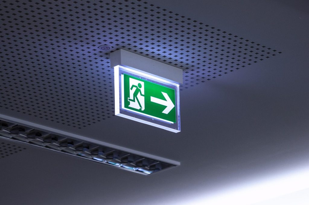 Illuminated emergency exit sign with a running figure and an arrow pointing right, mounted on a perforated ceiling in a commercial setting.