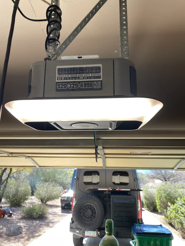 Close-up of a ceiling-mounted garage door opener by Unhinged Complete Door and Gate Service, with visible support brackets and wiring, and a van parked just outside the open garage door.