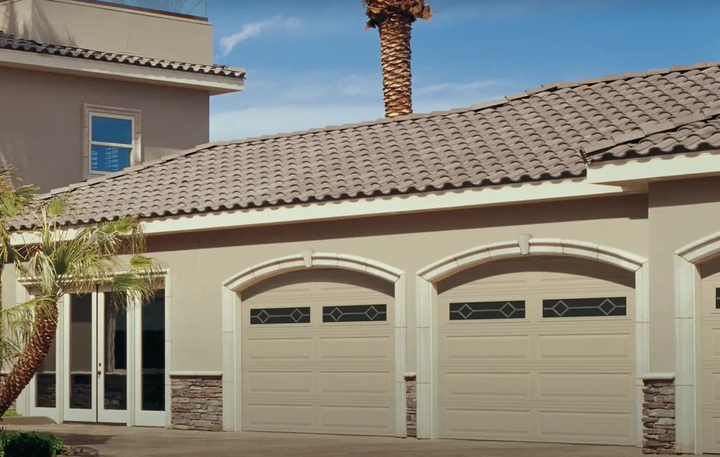 Modern beige home in Phoenix, Arizona, featuring three matching raised-panel garage doors with decorative windows, arched trim, and a tile roof—showcasing elegant curb appeal and desert-friendly design.