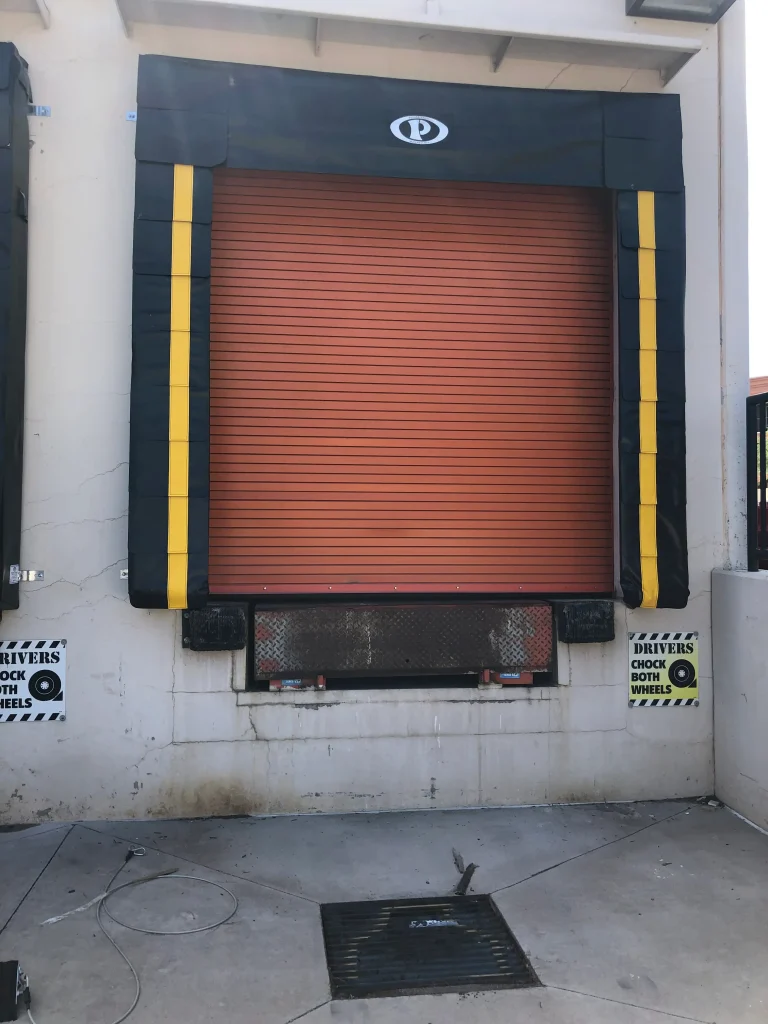 Closed red roll-up garage door at a commercial loading dock, framed with black and yellow protective bumpers, flanked by safety signs instructing drivers to chock both wheels.